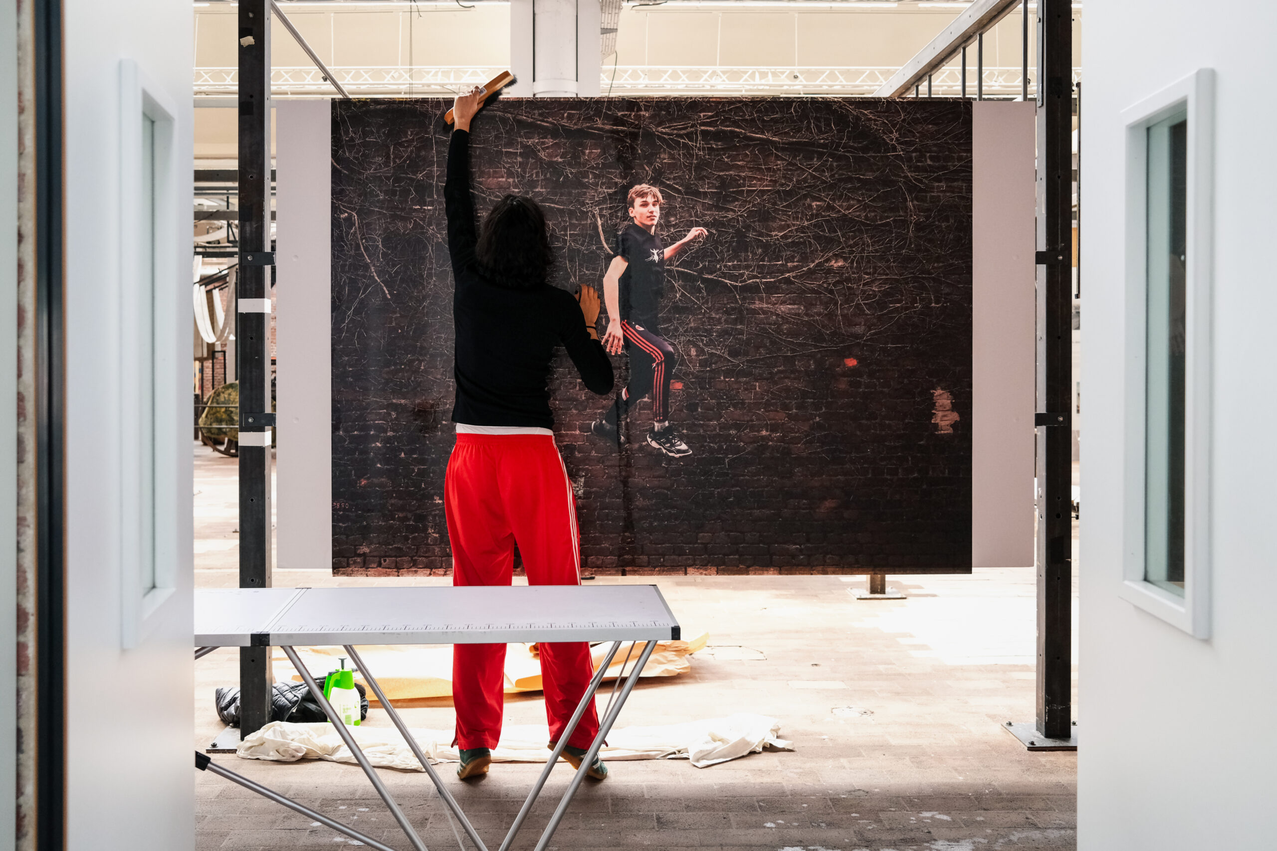 Une personne maroufle avec une brosse un grand tirage photographique collé sur un mur à l'intérieur d'une salle. L'image est celle de la photographe Anouk Desury dont le travail est visible dans l'exposition collective "Roubaix Sport" organisée par le Musée de La Manufacture, disponible du 20 août 27 octobre 2024 (vernissage prévu le 6 septembre).