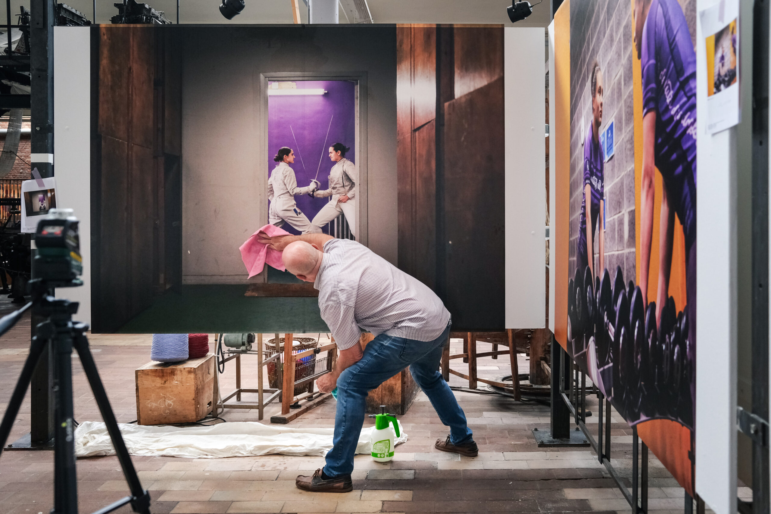 Un homme essuie un grand tirage photographique collé sur un mur à l'intérieur d'une salle. L'image est celle de la photographe Anouk Desury dont le travail est visible dans l'exposition collective "Roubaix Sport" organisée par le Musée de La Manufacture, disponible du 20 août 27 octobre 2024 (vernissage prévu le 6 septembre).