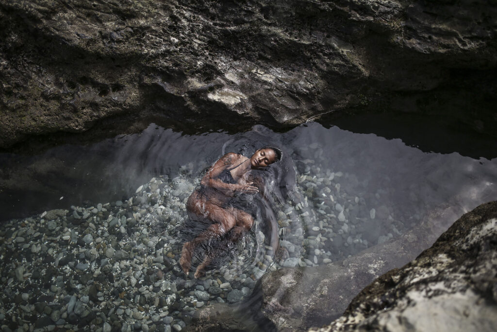 Une jeune femme en maillot de bain s'immerge dans de l'eau de mer entre des roches. Cette photographie est issue du long reportage réalisé en immersion par Gabriela Tellez (Light Motiv) sur les communautés des femmes surfeuses de l'Océan Pacifique à Nosara au Costa Rica.