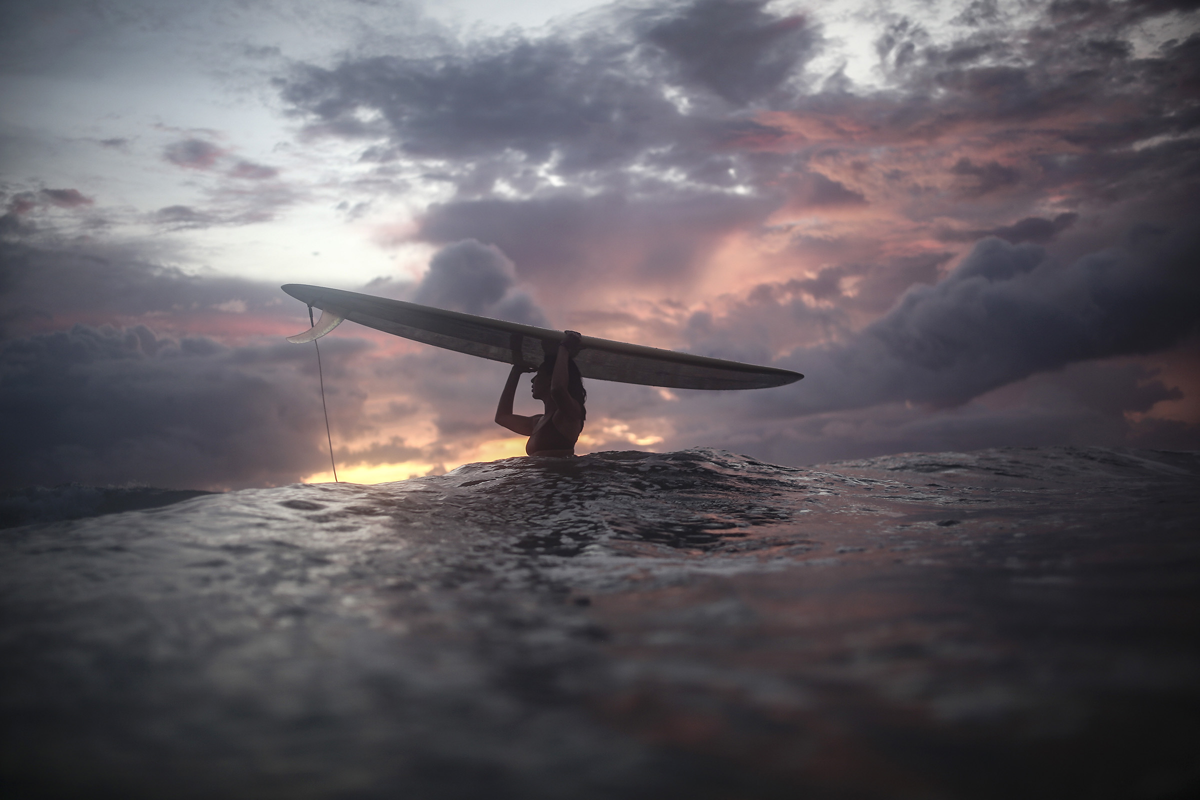 Une femme porte sa planche de surf au-dessus de sa tête, alors qu'elle se tient debout au milieu de l'eau. Cette photographie est issue du long reportage réalisé en immersion par Gabriela Tellez (Light Motiv) sur les communautés des femmes surfeuses de l'Océan Pacifique à Nosara au Costa Rica.