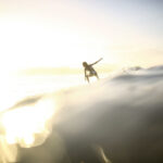 Une femme surfe sur une vague, la lumière du soleil l'éclairant entre contre-jour. Cette photographie est issue du long reportage réalisé en immersion par Gabriela Tellez (Light Motiv) sur les communautés des femmes surfeuses de l'Océan Pacifique à Nosara au Costa Rica.