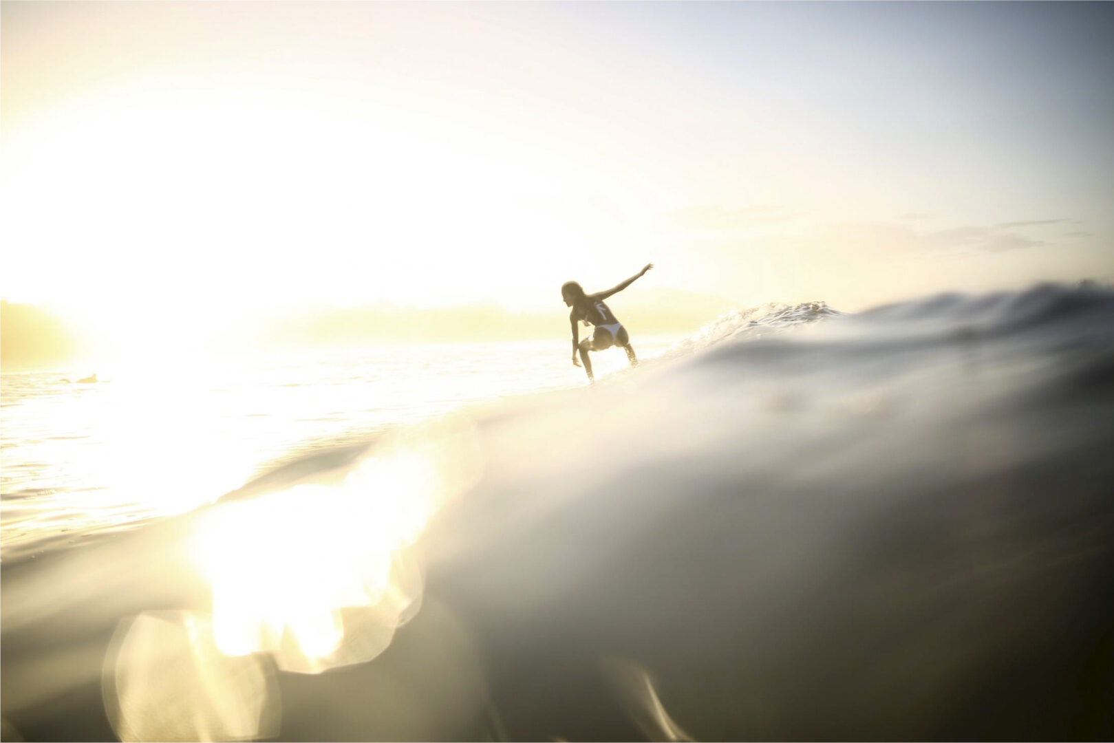 Une femme surfe sur une vague, la lumière du soleil l'éclairant entre contre-jour. Cette photographie est issue du long reportage réalisé en immersion par Gabriela Tellez (Light Motiv) sur les communautés des femmes surfeuses de l'Océan Pacifique à Nosara au Costa Rica.