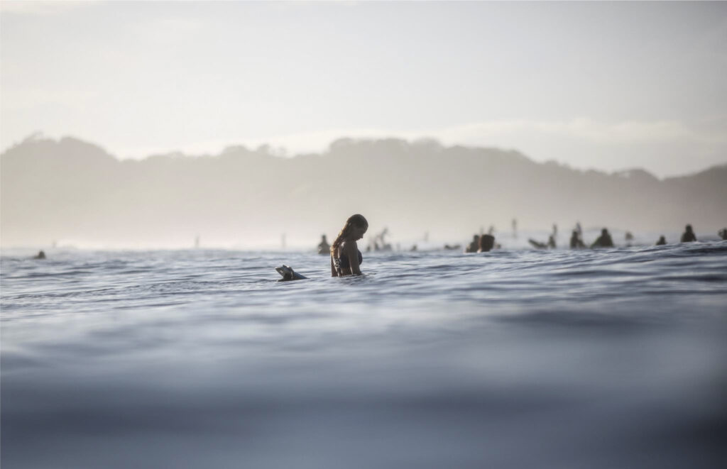 Une femme est visible assise sur sa planche au milieu de l'eau, avec en fond d'autres silhouettes de surfeuses. Cette photographie est issue du long reportage réalisé en immersion par Gabriela Tellez (Light Motiv) sur les communautés des femmes surfeuses de l'Océan Pacifique à Nosara au Costa Rica.