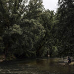 Une photographie d'une vue environnementale de la rivière de la Sambre avec un homme accroupi sur les berges, pensif. Fait partie de la série INLAND VOYAGE du photographe Quentin Pruvost dont le travail a été publié dans un livre aux Editions Light Motiv en 2022