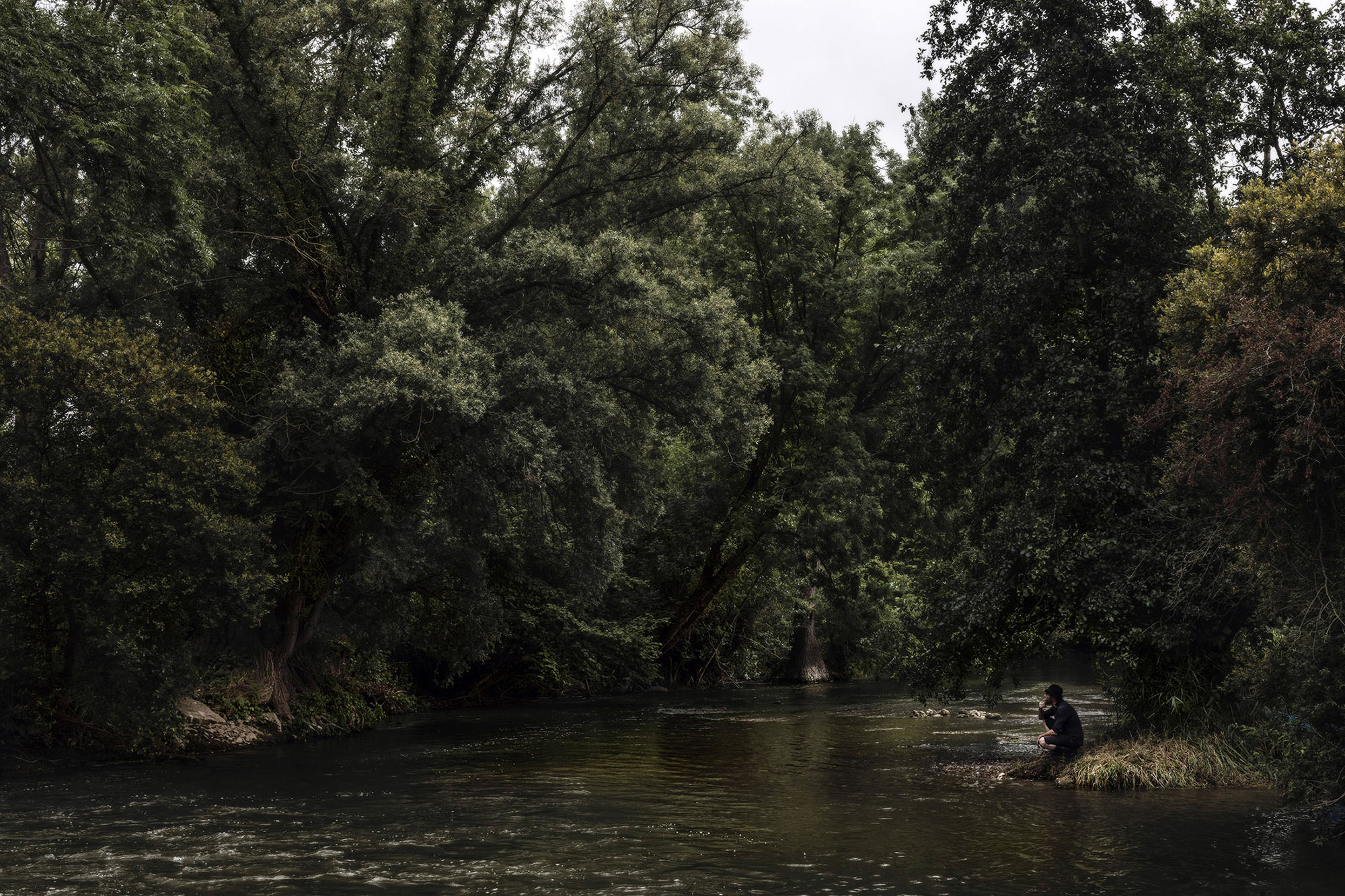 Une photographie d'une vue environnementale de la rivière de la Sambre avec un homme accroupi sur les berges, pensif. Fait partie de la série INLAND VOYAGE du photographe Quentin Pruvost dont le travail a été publié dans un livre aux Editions Light Motiv en 2022