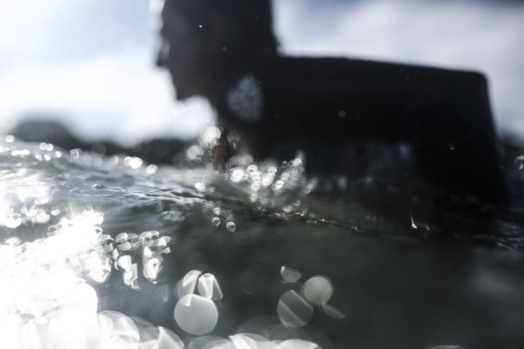 Une femme est visible en arrière-plan flouté, allongée sur son surf, pagayant pour avancer dans l'eau, alors que des gouttes de mer se dessinent en sphères de lumières en premier plan. Cette photographie est issue du long reportage réalisé en immersion par Gabriela Tellez (Light Motiv) sur les communautés des femmes surfeuses de l'Océan Pacifique à Nosara au Costa Rica.