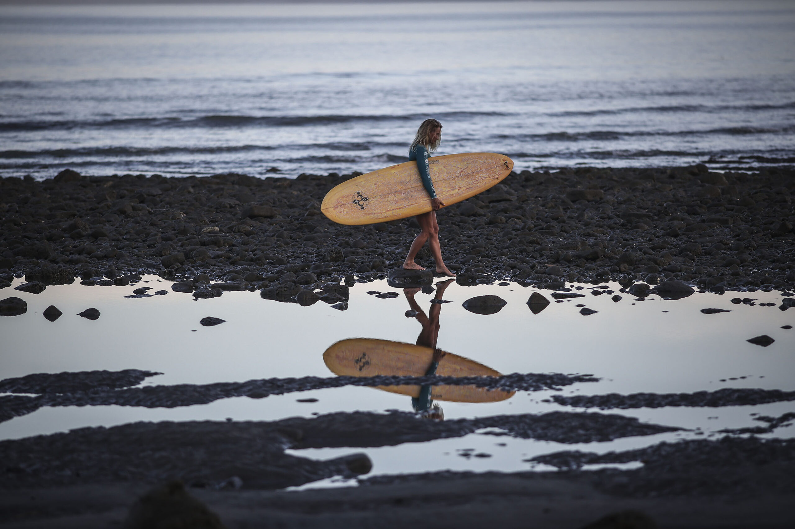 Une femme tenant une planche de surf sous le bras marche sur la plage de galets, l'océan en arrière-plan, et son reflet se répétant dans une flaque d'eau salée au premier plan. Cette photographie est issue du long reportage réalisé en immersion par Gabriela Tellez (Light Motiv) sur les communautés des femmes surfeuses de l'Océan Pacifique à Nosara au Costa Rica.