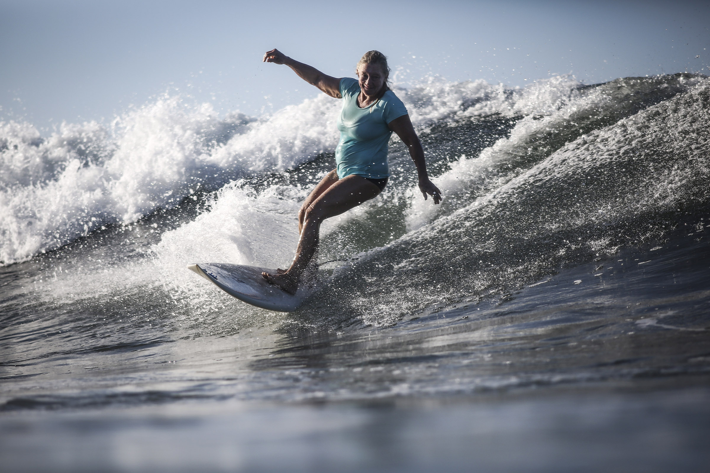 Une femme âgée en maillot de bain surfe sur une vague debout sur une planche. Cette photographie est issue du long reportage réalisé en immersion par Gabriela Tellez (Light Motiv) sur les communautés des femmes surfeuses de l'Océan Pacifique à Nosara au Costa Rica.