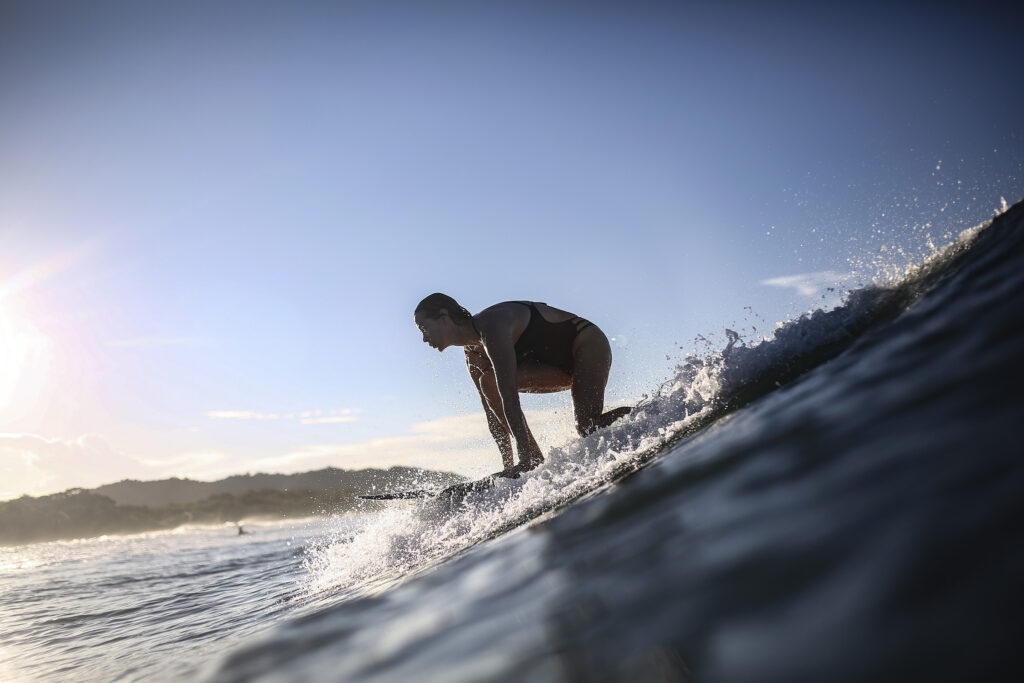 Une femme en maillot de bain s'apprête à se mettre debout sur sa planche pour surfer sur une vague. Cette photographie est issue du long reportage réalisé en immersion par Gabriela Tellez (Light Motiv) sur les communautés des femmes surfeuses de l'Océan Pacifique à Nosara au Costa Rica.