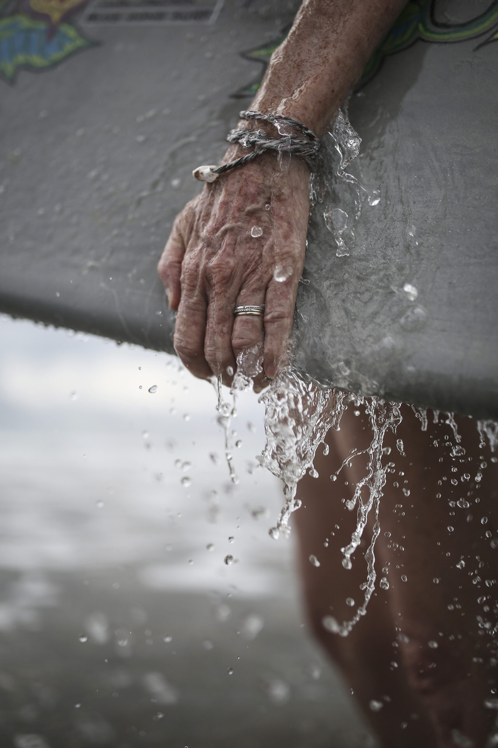 Une main de femme mouillée tient une planche de surf recouverte de gouttes d'eau. Cette photographie est issue du long reportage réalisé en immersion par Gabriela Tellez (Light Motiv) sur les communautés des femmes surfeuses de l'Océan Pacifique à Nosara au Costa Rica.